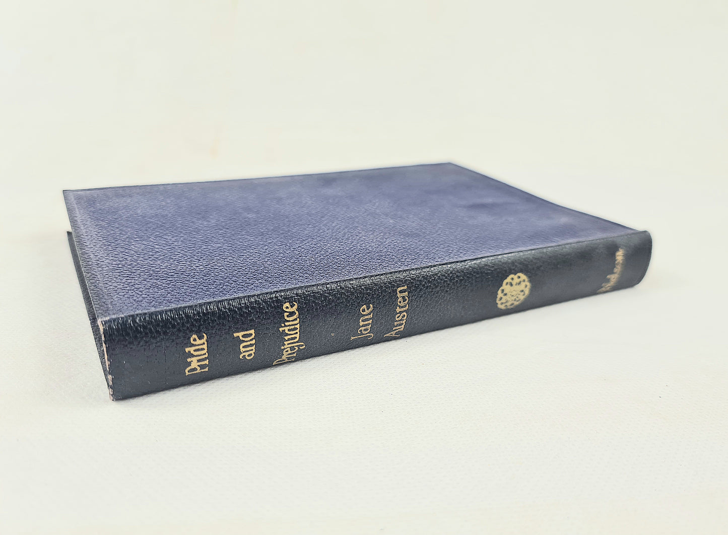 A vintage hardcover book, 'Pride and Prejudice' by Jane Austen, shown at an angle on a plain off-white background. The book features a textured navy blue cover with gold lettering on the spine and a gold decorative emblem.