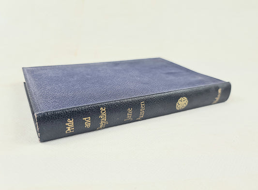 A vintage hardcover book, 'Pride and Prejudice' by Jane Austen, shown at an angle on a plain off-white background. The book features a textured navy blue cover with gold lettering on the spine and a gold decorative emblem.