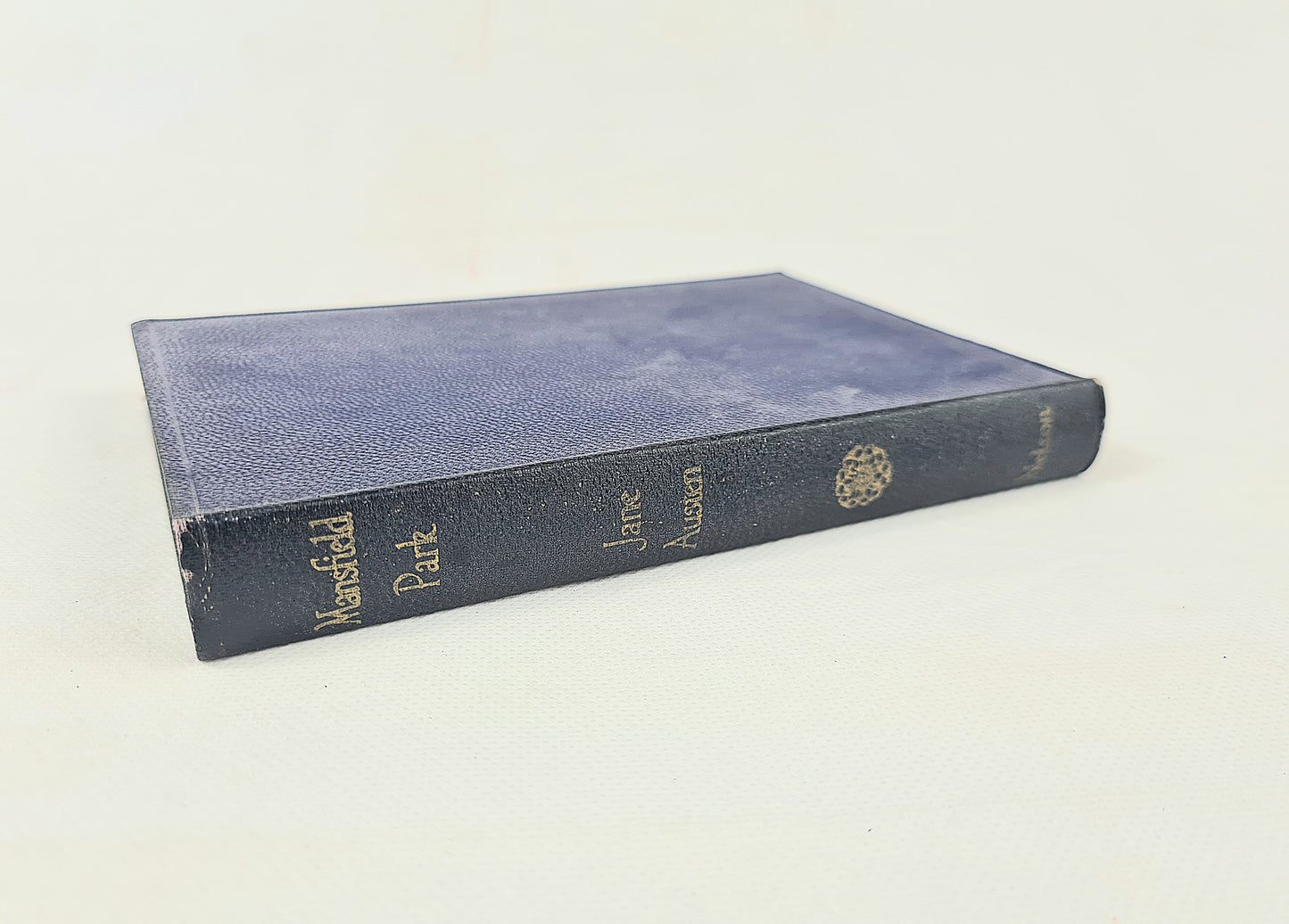 A vintage hardcover book titled 'Mansfeld Park' by Jane Austen, featuring a dark blue textured cover and gold lettering on the spine, photographed on a light off-white background.