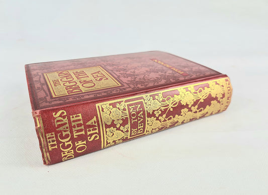 A vintage hardcover book, 'The Beggars of the sea by tom bevan, featuring a deep red marbled cover and ornate gold foil detailing on the spine and title block, photographed on a light off-white background.