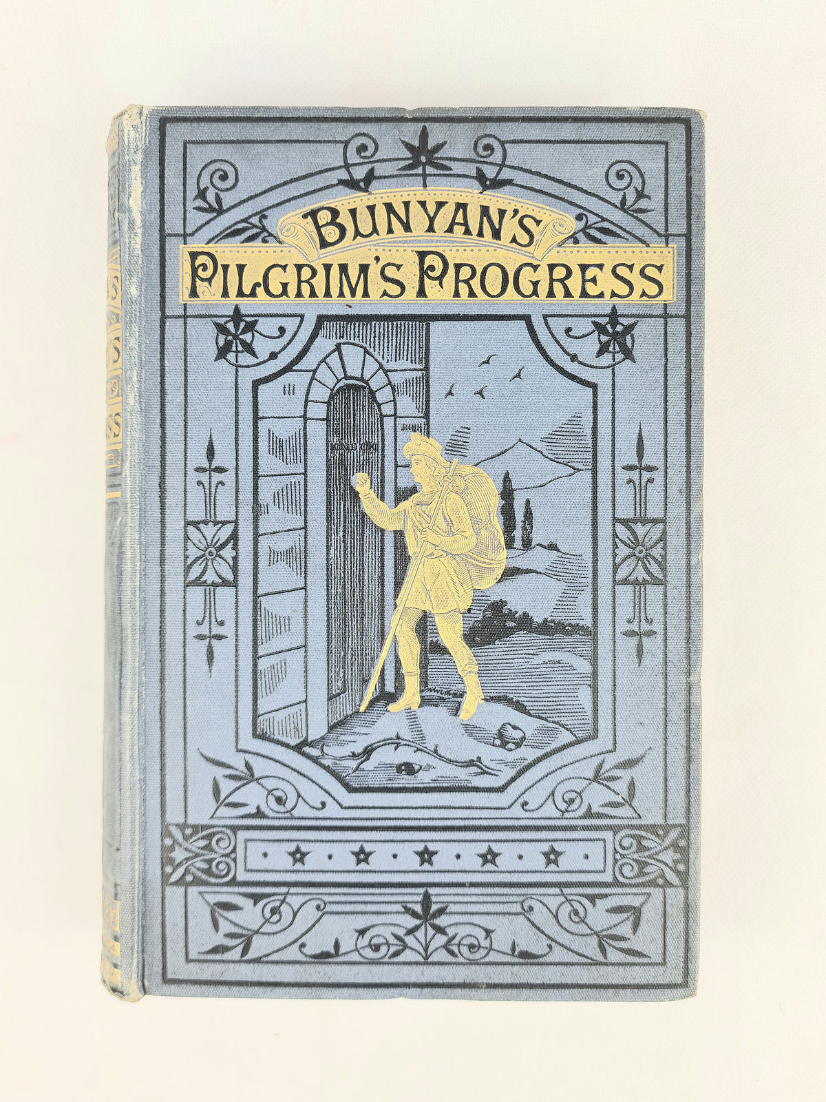 Vintage hardcover book of Bunyan's Pilgrim's Progress, featuring a light blue-gray illustrated cover with a gold-toned figure and title, photographed against a plain off-white background.