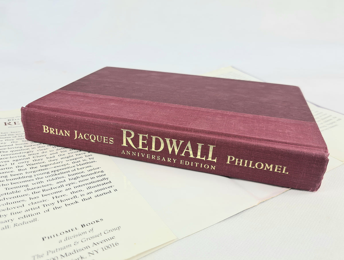 A hardcover book titled 'Redwall Anniversary Edition' by Brian Jacques, published by Philomel, resting on open book pages. The book cover is a deep burgundy red with gold lettering.